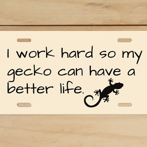 May include: A white license plate with black text that reads "I work hard so my gecko can have a better life." A black silhouette of a gecko is on the right side of the plate.