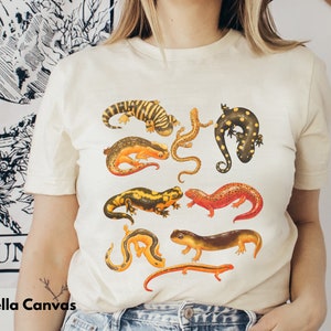 May include: A white t-shirt with a graphic print of various colorful salamanders. The salamanders are arranged in a random pattern on the shirt.