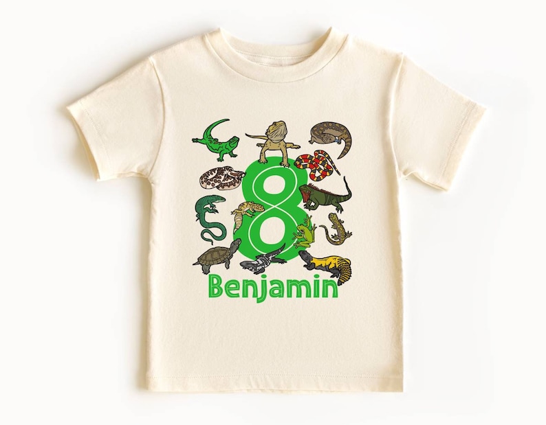 A light beige natural color kids shirt decorated with 13 reptile and amphibian characters with the number 8 in green. The name of a child is below the number. This would be a great personalizable birthday party shirt for children that love reptiles!