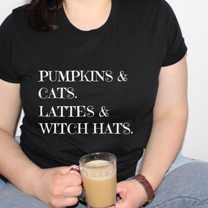 May include: A black t-shirt with white text that reads "Pumpkins & Cats. Lattes & Witch Hats."