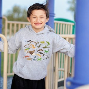 Puede incluir: Una persona joven lleva una sudadera gris con capucha con un estampado colorido de varios lagartos y serpientes. La sudadera tiene una capucha con cordón y un bolsillo canguro.