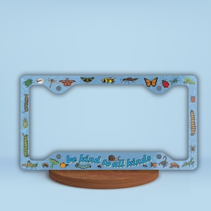 Puede incluir: Marco para matrícula azul con borde blanco y varias ilustraciones de insectos coloridos. El marco tiene el texto "be kind to all kinds" en letras azules.