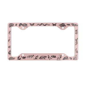 May include: A pink metal license plate frame with a black and white illustration of various animals around the border. The animals include a turtle, squirrel, snake, butterfly, deer, and more.