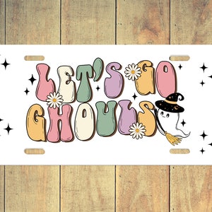Puede incluir: Una placa de matr&iacute;cula blanca con el texto "Let's Go Ghouls" en letras coloridas de estilo retro. Un fantasma de dibujos animados con un sombrero de bruja y una escoba vuela a la derecha del texto.