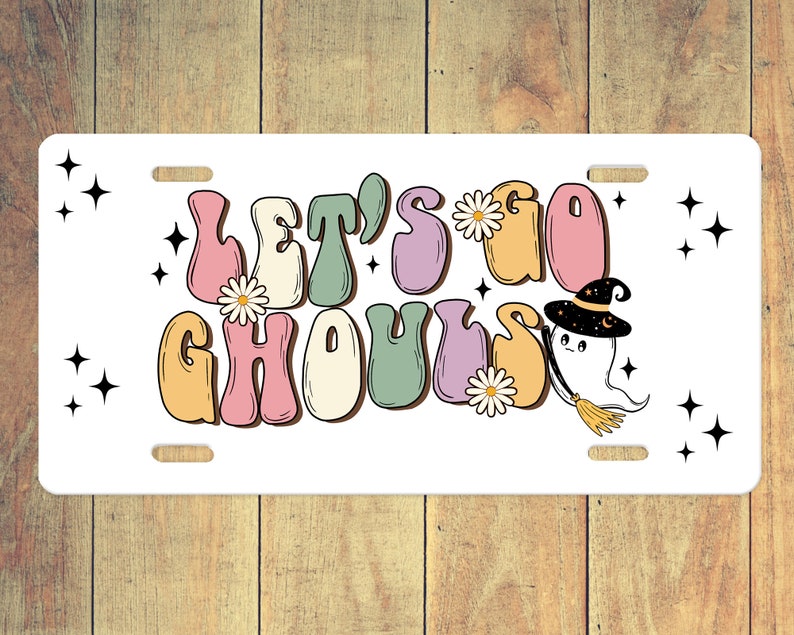 Puede incluir: Una placa de matr&iacute;cula blanca con el texto "Let's Go Ghouls" en letras coloridas de estilo retro. Un fantasma de dibujos animados con un sombrero de bruja y una escoba vuela a la derecha del texto.