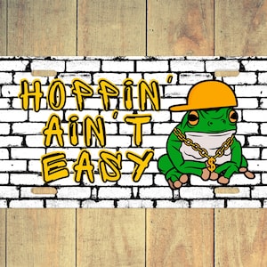 May include: A white brick wall with the words "Hoppin' Ain't Easy" in yellow graffiti. A green frog wearing a yellow hat and a gold chain with a dollar sign pendant sits in front of the wall.