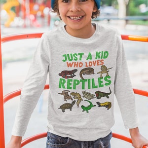 K&ouml;nnte beinhalten: Ein junges Kind tr&auml;gt ein graues Langarmshirt mit dem Text "Just a kid who loves reptiles" und verschiedenen bunten Reptilien-Illustrationen. Das Kind tr&auml;gt eine blaue M&uuml;tze und eine blaue Jeans.