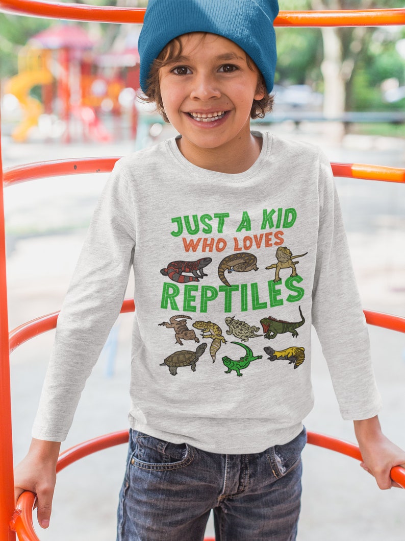 K&ouml;nnte beinhalten: Ein junges Kind tr&auml;gt ein graues Langarmshirt mit dem Text "Just a kid who loves reptiles" und verschiedenen bunten Reptilien-Illustrationen. Das Kind tr&auml;gt eine blaue M&uuml;tze und eine blaue Jeans.