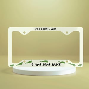 May include: White metal license plate frame with the text "FOR FROG'S SAKE" at the top and "GIMME SOME SPACE" at the bottom. Green frog silhouettes are printed along the bottom edge.