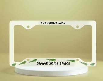 Könnte beinhalten: Weißer Nummernschildrahmen aus Metall mit dem Text "FOR FROG'S SAKE" oben und "GIMME SOME SPACE" unten. Grüne Froschsilhouetten sind am unteren Rand gedruckt.