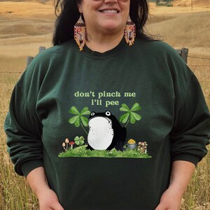 May include: A dark green sweatshirt with a black and white frog illustration and the text "don't pinch me I'll pee". The frog is surrounded by green shamrocks and gold coins.