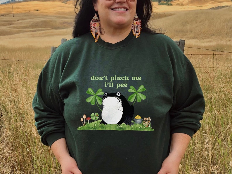 May include: A dark green sweatshirt with a black and white frog illustration and the text "don't pinch me I'll pee". The frog is surrounded by green shamrocks and gold coins.