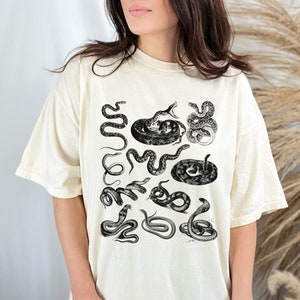 May include: A cream-coloured t-shirt with a black and white print of various snakes in different poses. The snakes are arranged in a random pattern on the shirt.