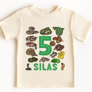 May include: Cream-colored t-shirt featuring a green number 5 and the name "SILAS" in green. The shirt is decorated with various cartoon snake illustrations in shades of brown, green, yellow, and red.