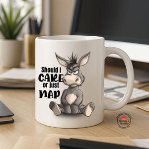 Könnte beinhalten: Eine weiße Keramik-Kaffeetasse mit einer Cartoon-Illustration eines Esels. Der Esel sitzt mit einem mürrischen Gesichtsausdruck und der Text "Should I CARE or just NAP" ist auf die Tasse gedruckt.