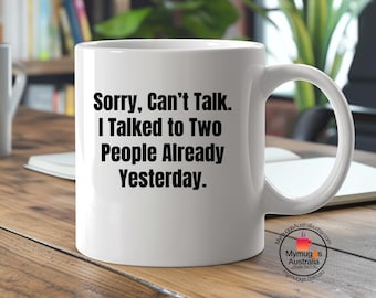 Könnte beinhalten: Weiße Keramik-Tasse mit dem Text "Sorry, Can't Talk. I Talked to Two People Already Yesterday." in Schwarz. Die Tasse hat einen gebogenen Henkel und steht auf einer Holzoberfläche.