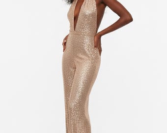 May include: A gold sequined jumpsuit with a halter neckline and wide-leg pants. The one-piece garment has a plunging neckline and is made of shimmering fabric. The model is standing with hands on hips, showcasing the outfit.