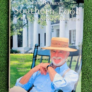 Puede incluir: La portada del libro de cocina "Southern Food & Plantation Houses" de Lee Bailey. El libro presenta a un hombre sonriente con sombrero de paja y camisa azul, sentado en una mecedora. El fondo muestra una casa de plantación blanca.