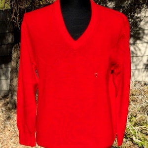 May include: A vibrant red V-neck sweater on a black mannequin. The long-sleeved jumper has a classic design and a small emblem near the chest. The background shows a blurred outdoor scene with trees and foliage.