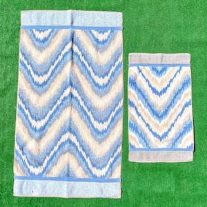May include: Two matching bath towels with a blue, beige, and white chevron pattern. The larger towel has a light blue border at the top and bottom. The smaller towel has a gray border. The towels are on a green surface.