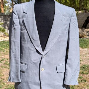 以下が含まれることがあります： シングルボタン留めと2つのフロントポケットが付いた、明るいグレーのブレザー。 ジャケットは黒いマネキンに表示されています。 ブレザーはノッチドラペルとクラシックなテーラードフィットです。 背景には緑と日光が写っています。