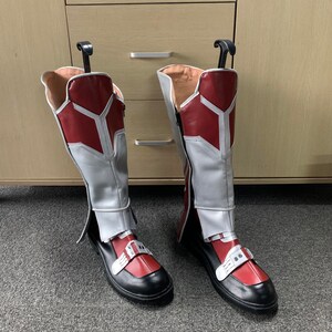 K&ouml;nnte beinhalten: Ein Paar rot-wei&szlig;e Stiefel mit schwarzen Sohlen. Die Stiefel haben einen Rei&szlig;verschluss an der Innenseite des Kn&ouml;chels und eine Schnalle auf dem Fu&szlig;r&uuml;cken.