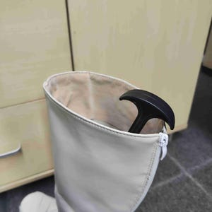 Op de afbeelding: Een hoge, gebroken witte laars met een ritssluiting. De binnenkant van de laars is lichtbeige. Een zwarte laarstrekker zit in de laars. De laars is gemaakt van een glad materiaal.