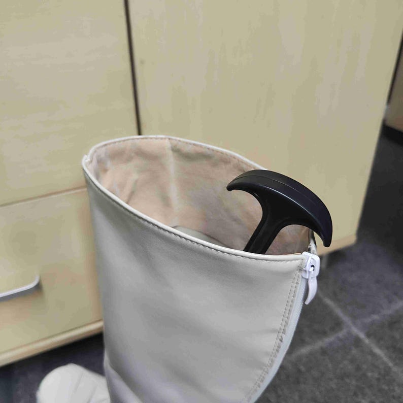 Op de afbeelding: Een hoge, gebroken witte laars met een ritssluiting. De binnenkant van de laars is lichtbeige. Een zwarte laarstrekker zit in de laars. De laars is gemaakt van een glad materiaal.