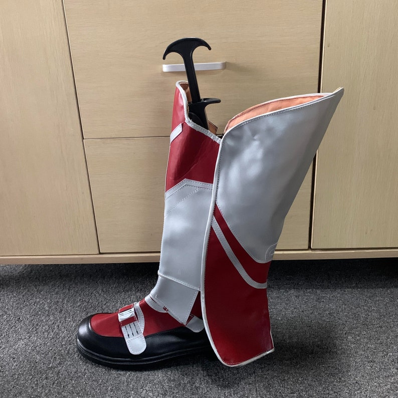 K&ouml;nnte beinhalten: Rote und wei&szlig;e kniehohe Stiefel mit einer schwarzen Schnalle. Die Stiefel haben ein wei&szlig;es Leder-Oberteil und einen roten Leder-Unterteil. Die Stiefel stehen auf einer Holzoberfl&auml;che.