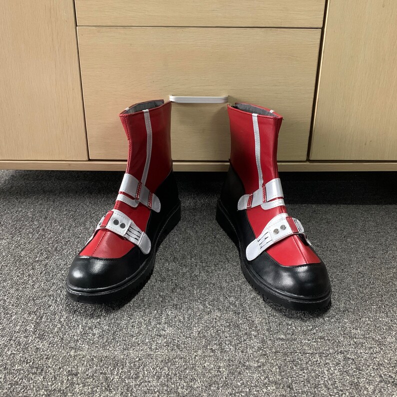 K&ouml;nnte beinhalten: Ein Paar rot-schwarze Stiefel mit wei&szlig;en Akzenten. Die Stiefel haben einen Schnallenriemen und eine spitze Zehenkappe.