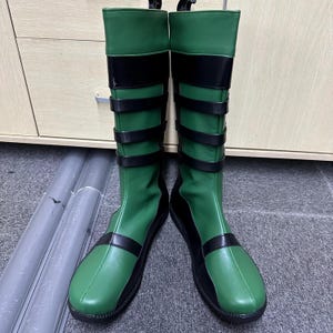 Op de afbeelding: Een paar groene en zwarte kniehoge laarzen. De laarzen hebben een groen bovenwerk met zwarte accenten, waaronder bandjes en een band rond de bovenkant. De zolen zijn zwart.