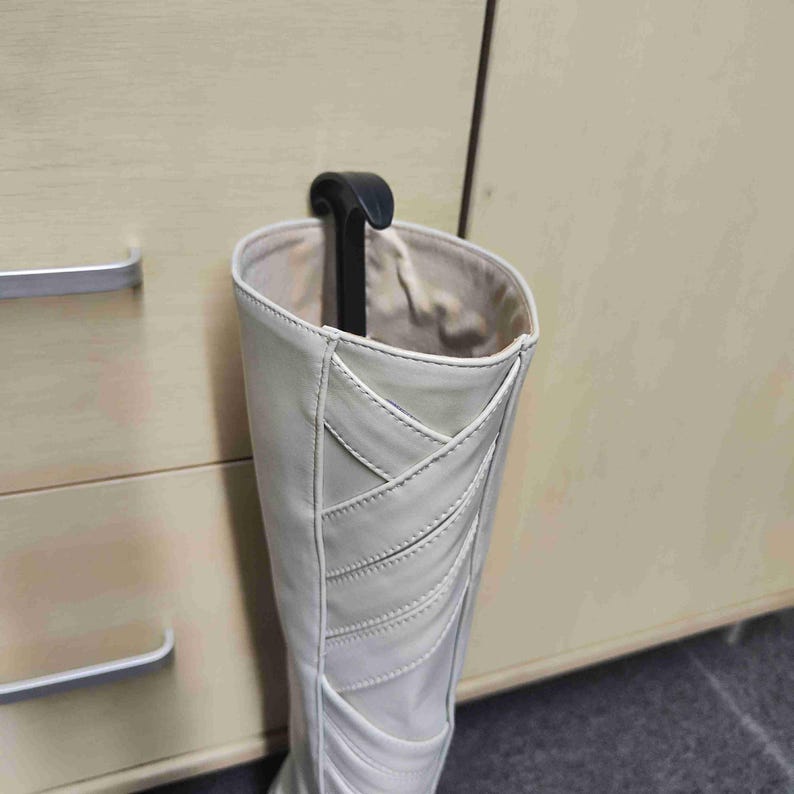 Op de afbeelding: Een hoge, gebroken witte laars met decoratieve stiksels wordt vastgehouden door een zwarte laarshanger. De laars is gemaakt van een glad materiaal en heeft een lichtgekleurde binnenkant. De hanger is bevestigd aan een houten kast.