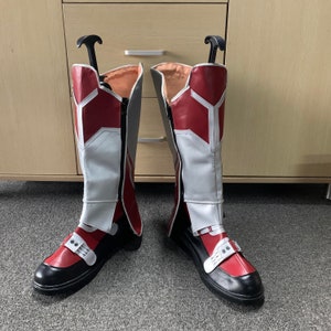 K&ouml;nnte beinhalten: Ein Paar rot-wei&szlig;e Stiefel mit schwarzen Sohlen. Die Stiefel haben einen Rei&szlig;verschluss an der Innenseite des Beins und einen Riemen &uuml;ber dem Fu&szlig;r&uuml;cken.