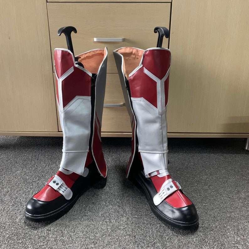 K&ouml;nnte beinhalten: Ein Paar rot-wei&szlig;e Stiefel mit schwarzen Sohlen. Die Stiefel haben einen Rei&szlig;verschluss an der Innenseite des Beins und einen Riemen &uuml;ber dem Fu&szlig;r&uuml;cken.