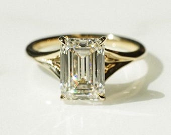 May include: A gold ring with an emerald-cut diamond. The diamond is the focal point, set in a classic four-prong setting. The band is a simple, polished gold. The ring is displayed on a white surface, with soft lighting.