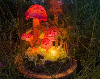 May include: A glass dome with a wooden base containing a miniature forest scene with glowing red mushrooms and a small white picket fence. The scene is lit by a small yellow light.