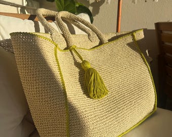 May include: A large, beige woven tote bag with a lime green trim and a matching tassel. The bag has two sturdy handles and is sitting on a white surface. The bag is made of a natural fiber material.