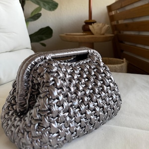 May include: A silver-colored, woven clutch bag with a structured frame and a top handle. The bag has a textured, basket-weave appearance. The background includes a white pillow, a wooden table, and a decorative lamp.