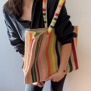Puede incluir: Un bolso tote de crochet a rayas verticales de colores. El bolso presenta rayas rosas, amarillas, naranjas, verdes y azules. Las correas tambi&eacute;n est&aacute;n tejidas con un patr&oacute;n de colores repetido. El bolso lo sostiene una persona.