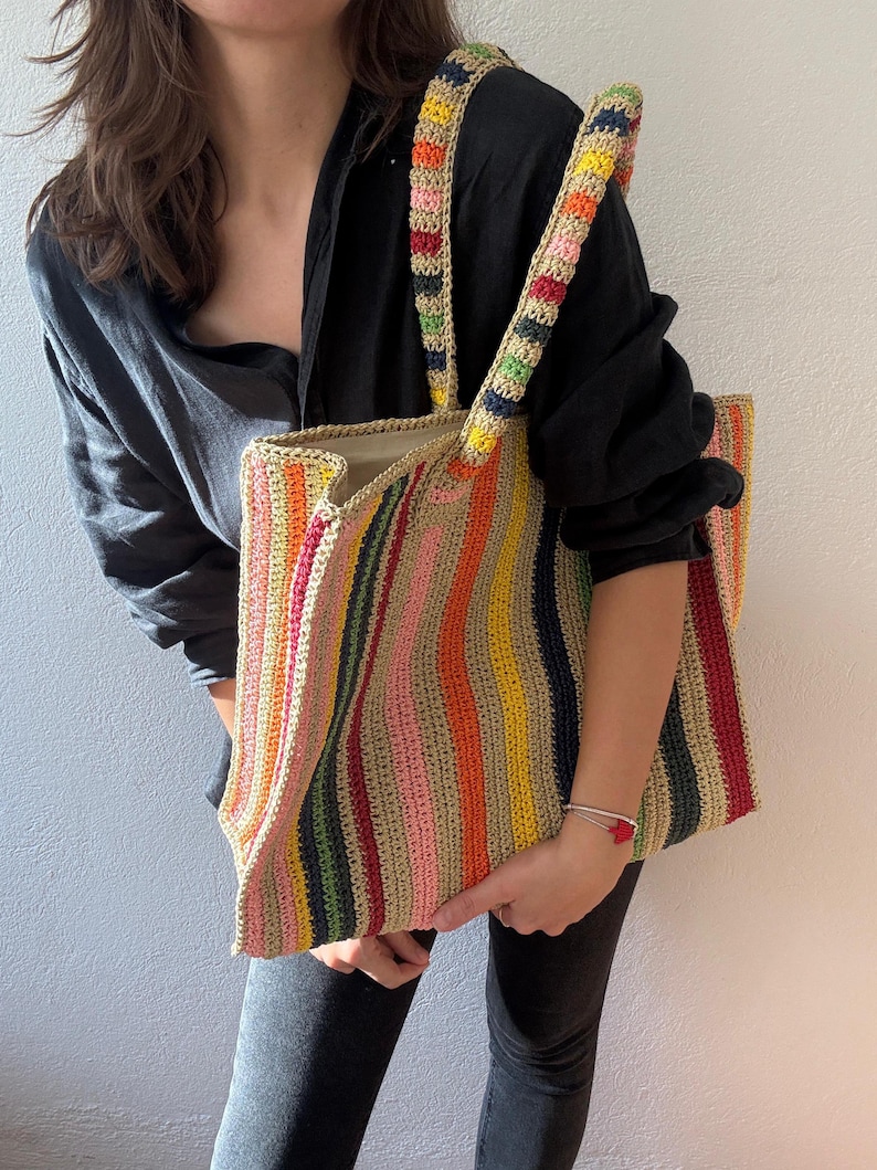 Puede incluir: Un bolso tote de crochet a rayas verticales de colores. El bolso presenta rayas rosas, amarillas, naranjas, verdes y azules. Las correas tambi&eacute;n est&aacute;n tejidas con un patr&oacute;n de colores repetido. El bolso lo sostiene una persona.