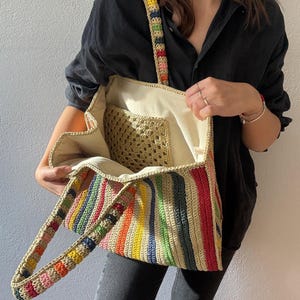 Puede incluir: Un bolso tote de crochet hecho a mano con un patr&oacute;n de rayas arco&iacute;ris. El bolso presenta un interior beige y un bolsillo de crochet m&aacute;s peque&ntilde;o. Las asas y la correa est&aacute;n adornadas con un dise&ntilde;o vibrante y multicolor. Este elegante bolso es perfecto para llevar lo esencial del d&iacute;a a d&iacute;a.