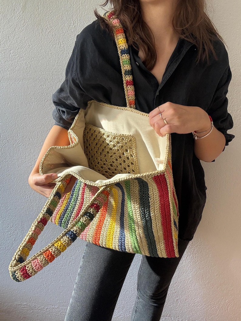 Puede incluir: Un bolso tote de crochet hecho a mano con un patr&oacute;n de rayas arco&iacute;ris. El bolso presenta un interior beige y un bolsillo de crochet m&aacute;s peque&ntilde;o. Las asas y la correa est&aacute;n adornadas con un dise&ntilde;o vibrante y multicolor. Este elegante bolso es perfecto para llevar lo esencial del d&iacute;a a d&iacute;a.