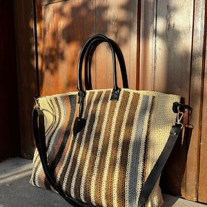 May include: A striped crocheted tote bag with black leather handles and a long strap. The bag is made of natural fibers and has a casual, relaxed style.