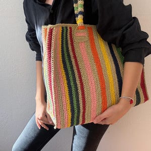 Puede incluir: Un bolso tote de crochet a rayas de colores con un asa a juego. El bolso presenta rayas verticales en varios colores, incluyendo rojo, naranja, amarillo, verde, rosa y azul. El bolso es sostenido por una persona que lleva una camisa negra y vaqueros.