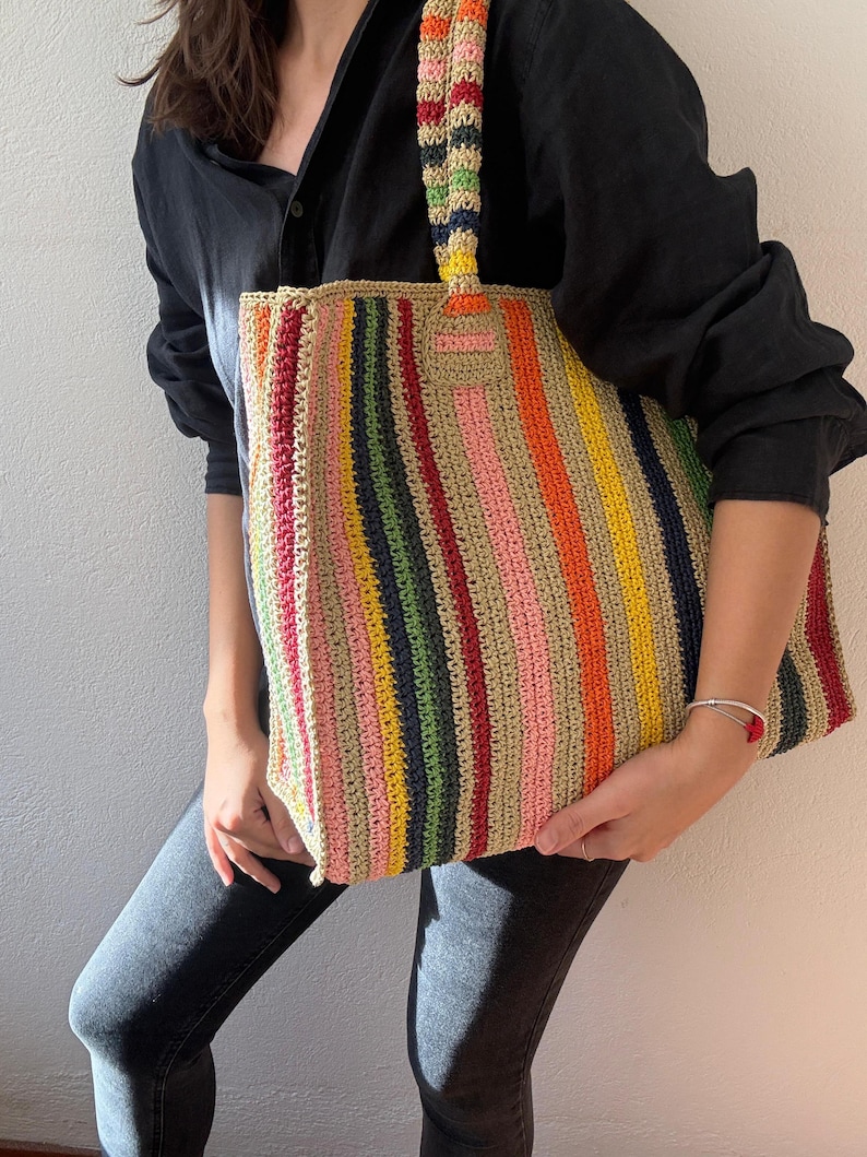 Puede incluir: Un bolso tote de crochet a rayas de colores con un asa a juego. El bolso presenta rayas verticales en varios colores, incluyendo rojo, naranja, amarillo, verde, rosa y azul. El bolso es sostenido por una persona que lleva una camisa negra y vaqueros.
