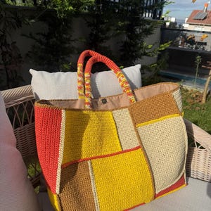 May include: A large, patchwork crochet tote bag with a variety of colors including red, yellow, brown, and cream. The bag has two orange and yellow handles. The bag is sitting on a wicker chair.