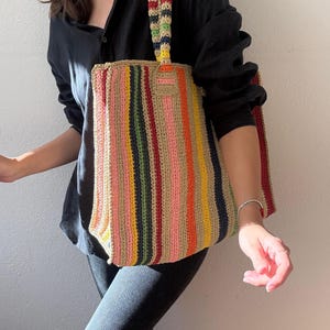 Puede incluir: Bolso tote de crochet hecho a mano con rayas verticales multicolores, incluyendo rosa, amarillo, verde y marr&oacute;n. El bolso tiene un asa a rayas a juego y una forma rectangular, perfecto para llevar objetos.