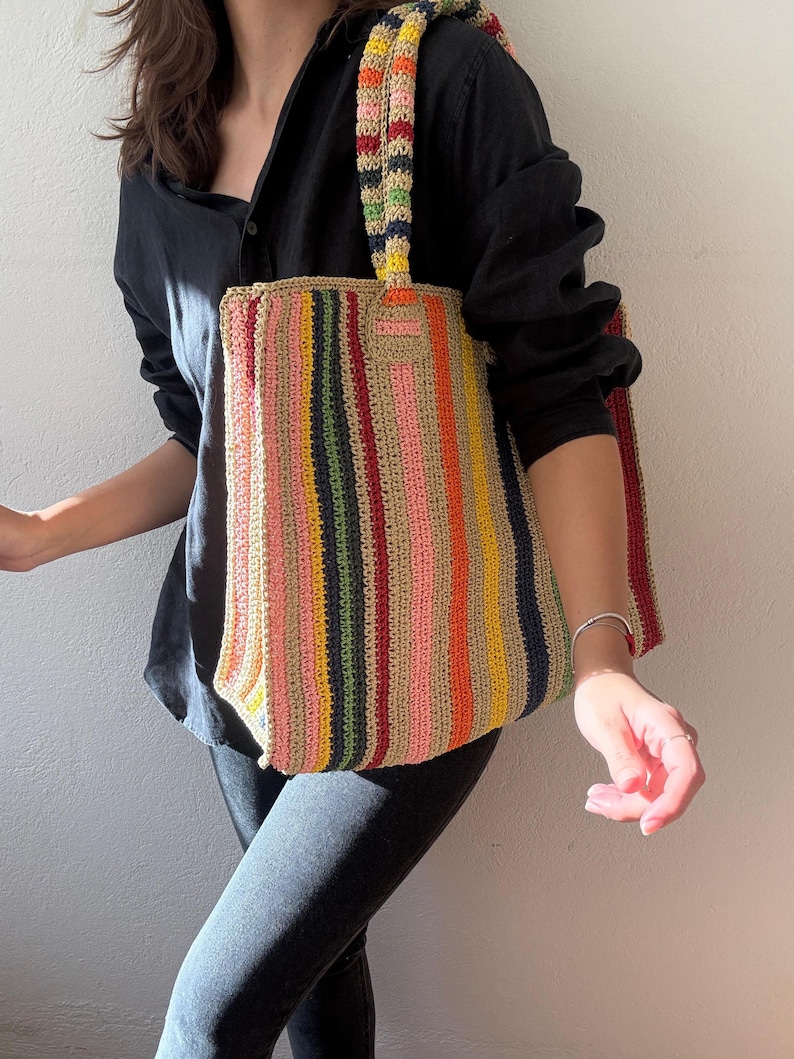 Puede incluir: Bolso tote de crochet hecho a mano con rayas verticales multicolores, incluyendo rosa, amarillo, verde y marr&oacute;n. El bolso tiene un asa a rayas a juego y una forma rectangular, perfecto para llevar objetos.