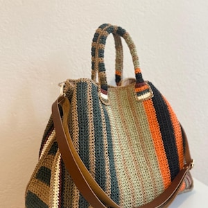 Puede incluir: Bolso tote de crochet a rayas en tonos marrón, verde, naranja y negro. Cuenta con una correa de hombro de cuero marrón y asas de crochet a juego. Un accesorio hecho a mano. Mide aproximadamente 40 cm de ancho y 30 cm de alto.