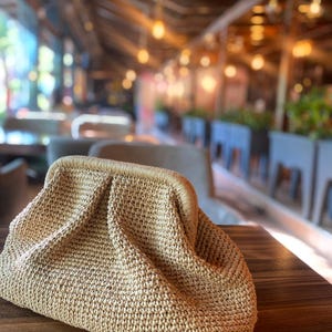 May include: A beige, woven clutch bag with a gathered top and a structured frame. The bag is made of a natural fiber material and sits on a dark wooden surface. The background is blurred, suggesting an outdoor setting.